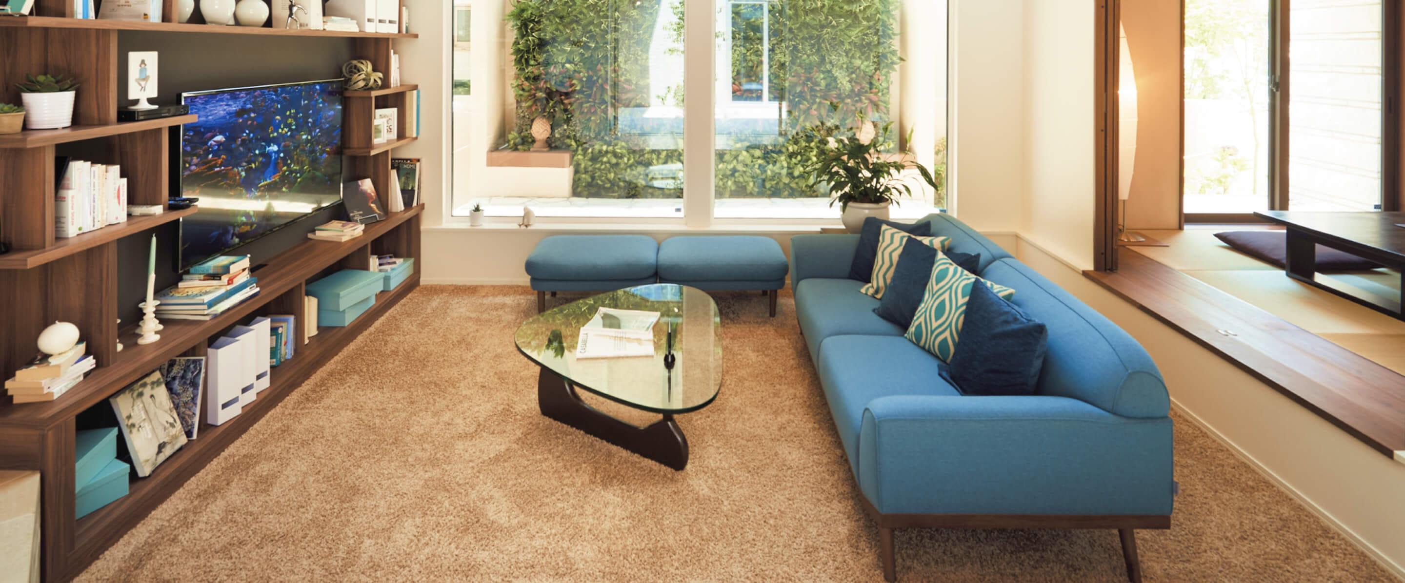 明るいリビングルームのインテリア。青いソファとガラスのローテーブル、木製の本棚や窓際の観葉植物が配置され、ナチュラルな雰囲気の住まい。